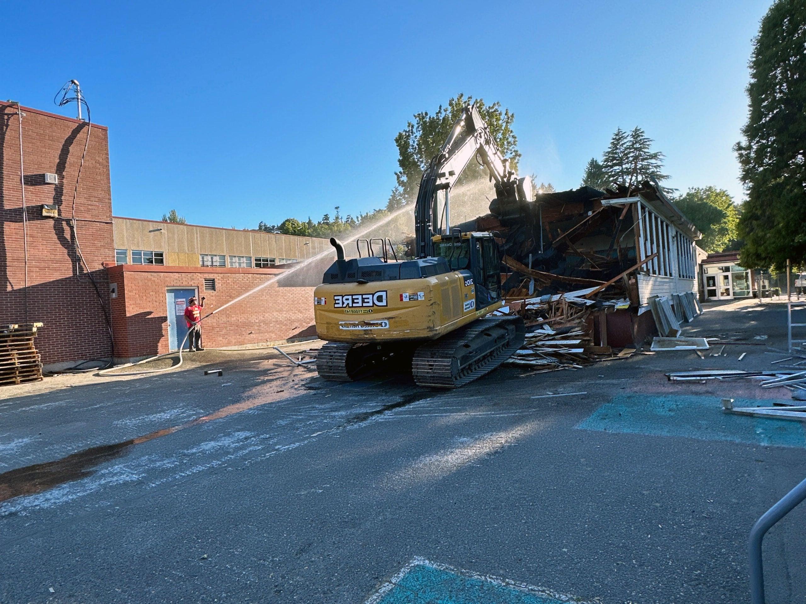 an excavator is crushing a wooden building while a person sprays a hose at it