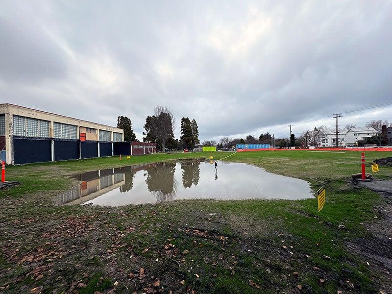 a pond of water sites amid grass near a 2-story building. there is an orange construction fence visible.