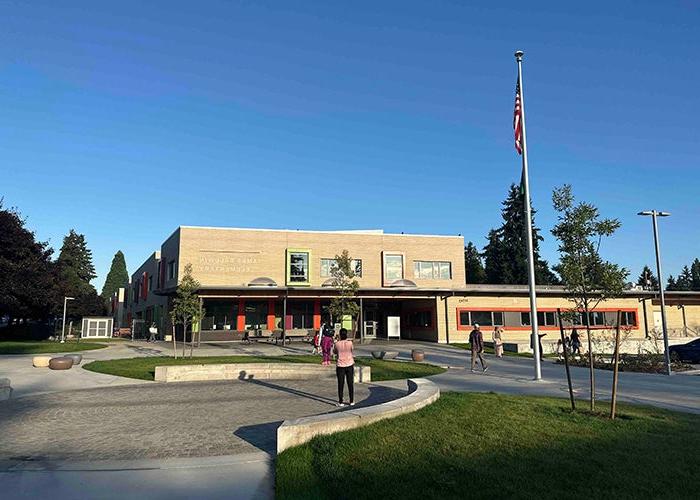 the front of a building has one part that is 2-stories and 1 part that is 1-story. it is light colored with a roofed entry. a circular paved area, an area of grass, a concrete walkway, and a flag pole are in front. there are a few people facing away from the camera