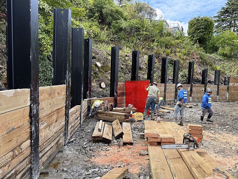 large metal posts approximately 12 feet tall are lined up along a hillside and workers are putting giant wood slabs between them